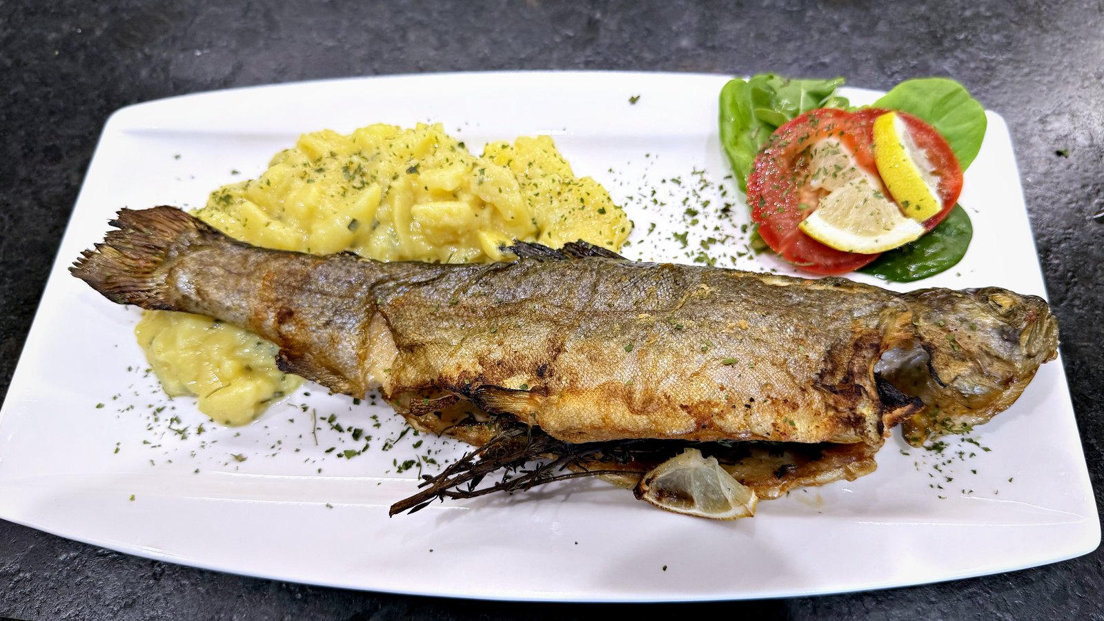 Ein weißer Teller mit einer ganzen gebratenen Forelle mit goldbrauner Haut auf Erdäpfelsalat, daneben zwei Paradeiserscheiben mit einer Zitronenspalte auf einem Salatblatt.